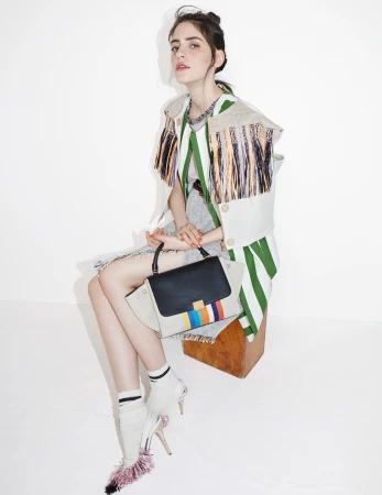 A woman in a striped jacket sits on a wooden stool, holding a color-blocked handbag, while wearing white high heel shoes with pink trim.