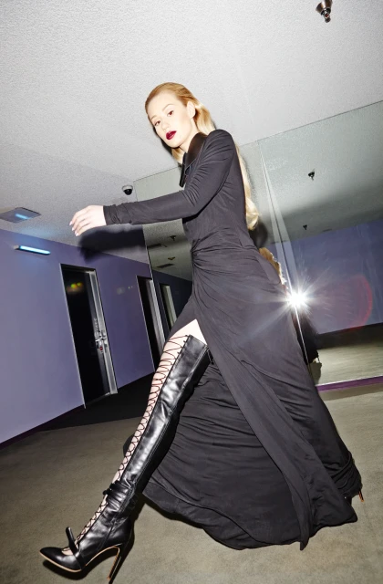 Fashion portrait of a blonde woman in a long black dress and lace-up boots walking toward the camera in an indoor setting.