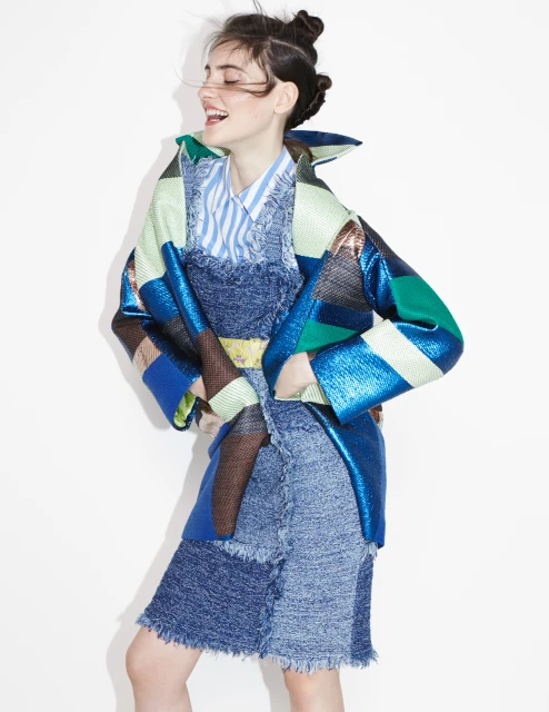 Smiling woman in a blue patchwork dress and a shimmering blue, green, and gold jacket posing against a plain white backdrop in a studio.