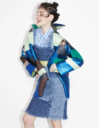 Smiling model with double hair buns wearing a blue patterned coat over a striped shirt against a plain white background