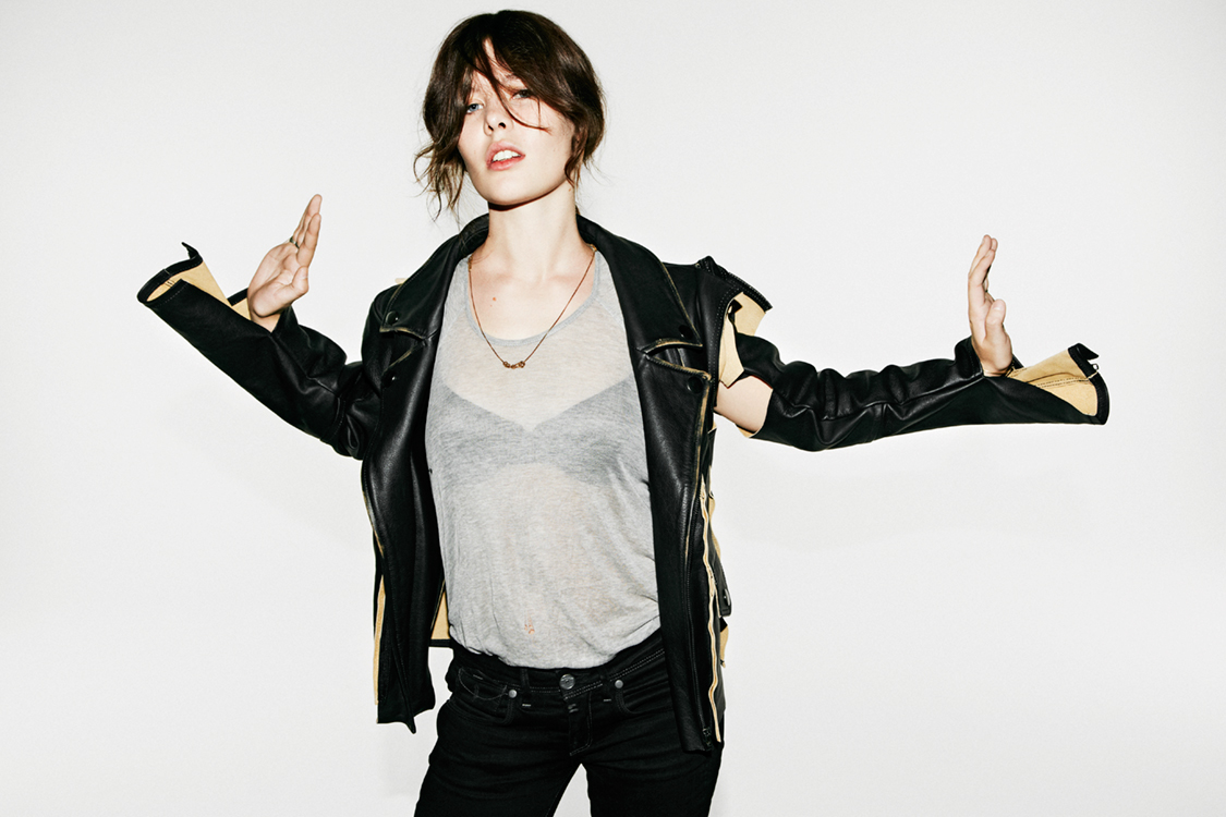 Woman wearing a black leather jacket over a grey t-shirt holding her hands up against a white background