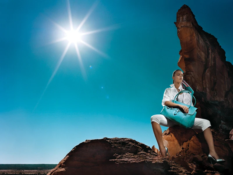 Adult woman sitting on rocks holding a teal purse, wearing white clothing, teal high heels, clear sky, and desert landscape.