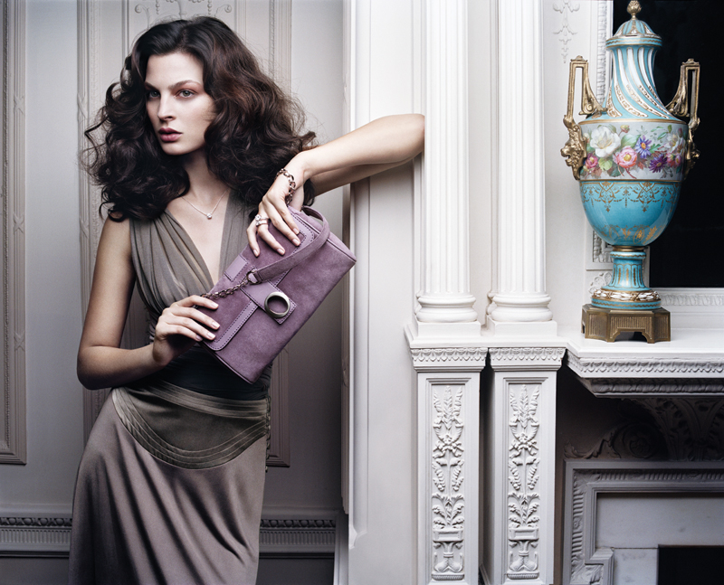 Adult female model with curly dark hair, wearing a taupe dress and holding a lilac-colored purse, posing next to a decorative urn.