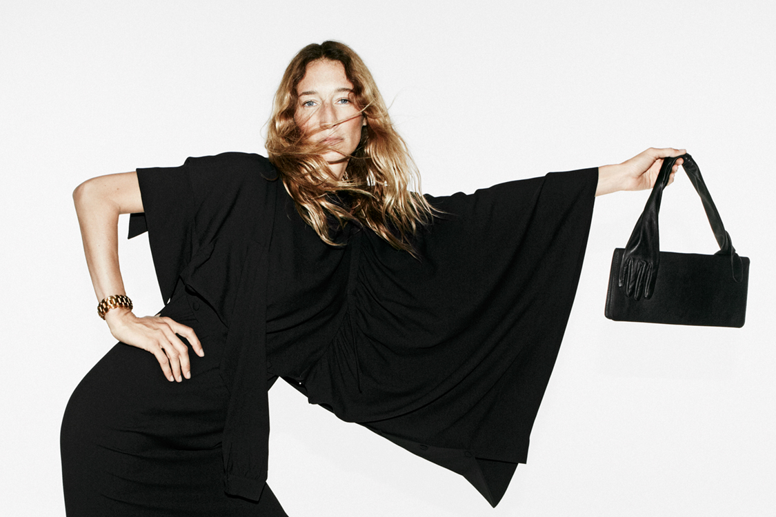 Blonde woman poses in a black dress with a purse against a white backdrop, accented by a gold bracelet and dynamic hair movement.