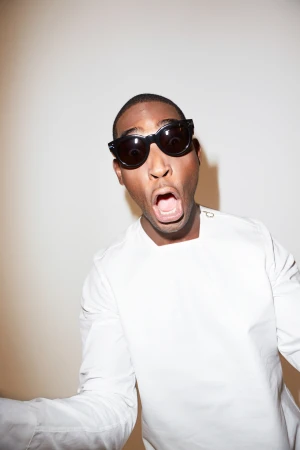 Close-up of an adult male with open mouth, wearing sunglasses and a white shirt against a light background, looking at the camera.