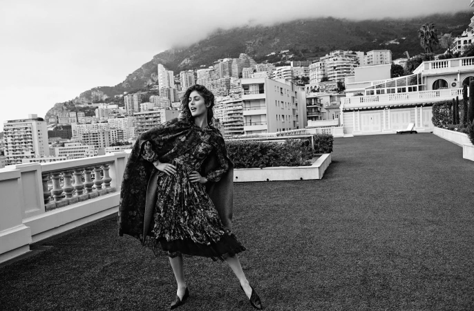 Monochrome fashion portrait of a smiling woman posing in a patterned dress on a rooftop overlooking a coastal city.