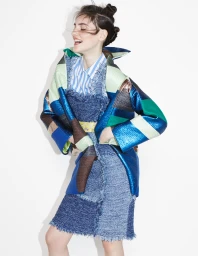 Smiling model with double hair buns wearing a blue patterned coat over a striped shirt against a plain white background