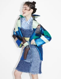 Smiling woman wearing a blue patterned jacket over a striped shirt and denim dress against a white background