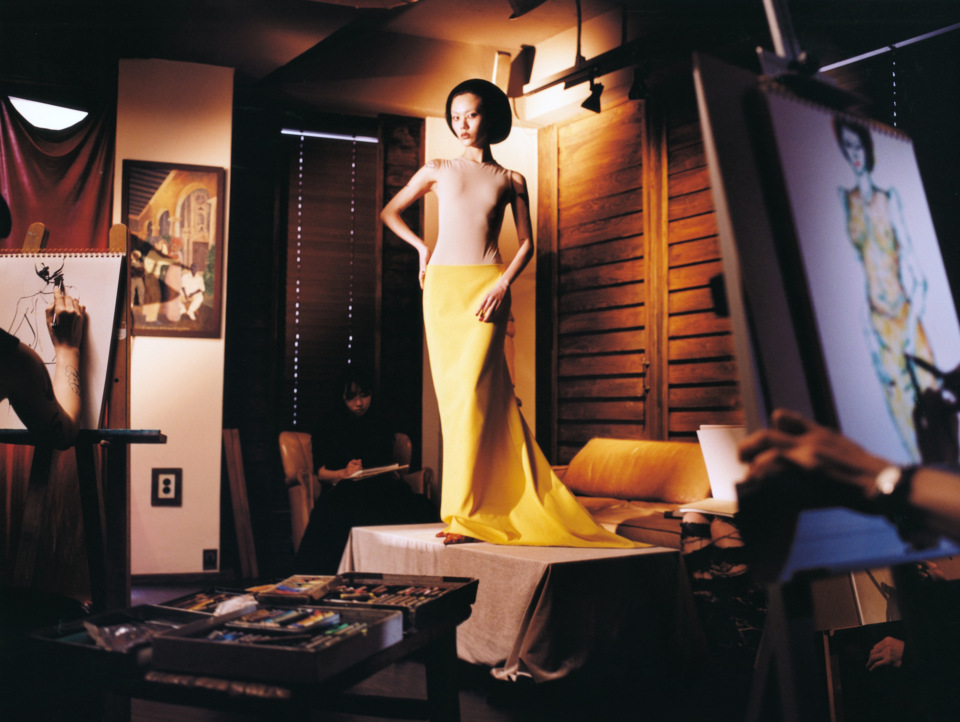 A slender woman poses in a yellow gown in an artist's studio, surrounded by canvases, paints, and artists at work in a warm, creative indoor space.