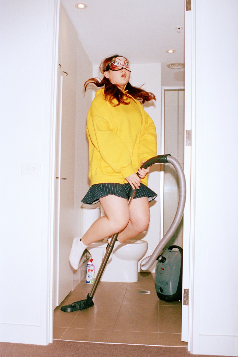 Woman in yellow hoodie levitates with a vacuum cleaner in a bathroom, wearing a sleep mask and white sneakers with a playful expression.