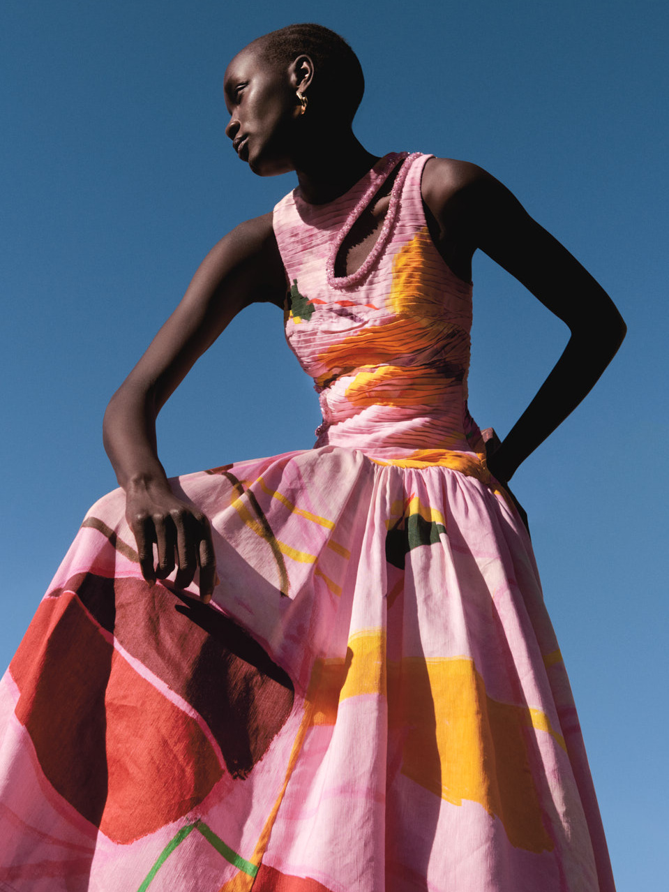 Fashion shot featuring a Black woman in a pink dress with abstract patterns, posing against a clear blue sky in natural light.