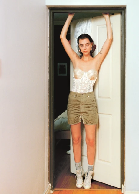 A woman poses in a doorway wearing a corset and mini skirt with white high-top sneakers against a residential interior backdrop.