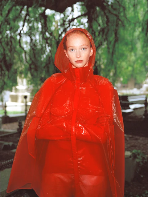 Red-haired woman in a bright red, transparent hooded raincoat stands in a garden with trees and headstones in the background.