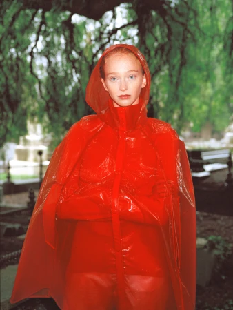 Red-haired woman in a bright red, transparent hooded raincoat stands in a garden with trees and headstones in the background.