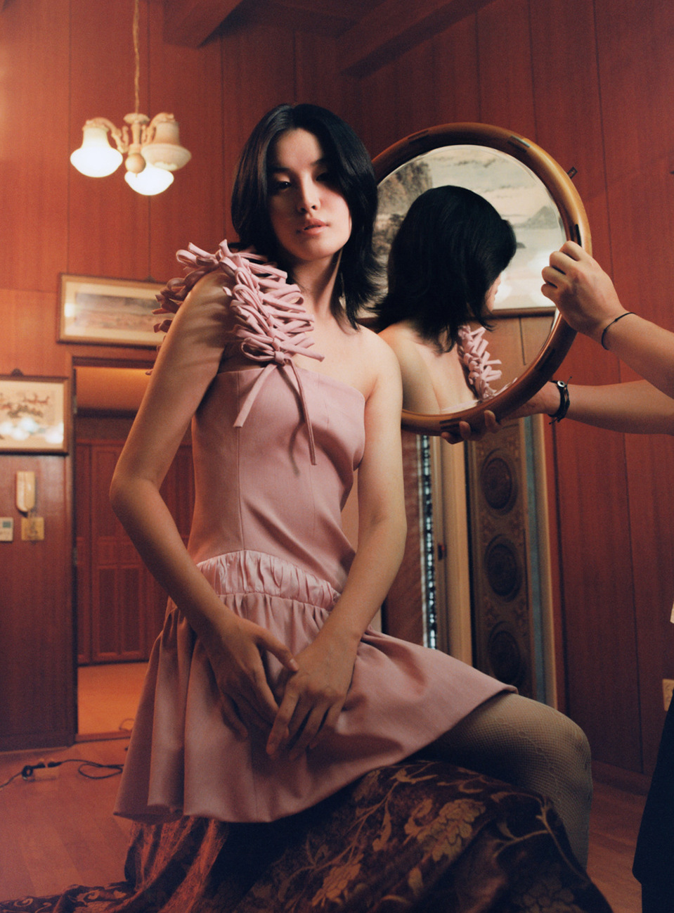 A seated woman in a pink dress glances towards her mirror reflection, held by hands off-camera, in a vintage interior setting.