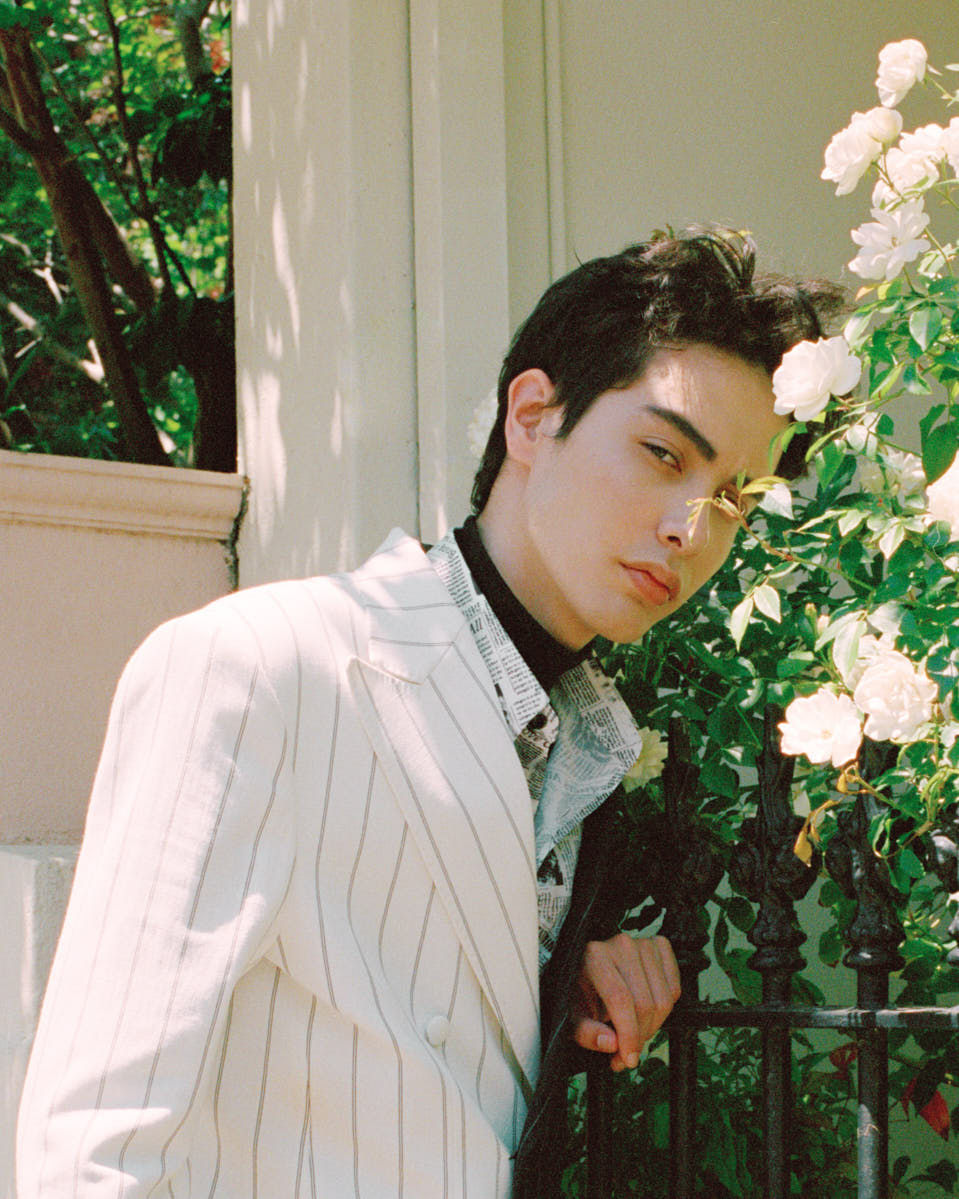 Fashion portrait of a young adult male in a pinstripe suit, posing outdoors near a wrought iron fence and white roses, shallow depth of field.