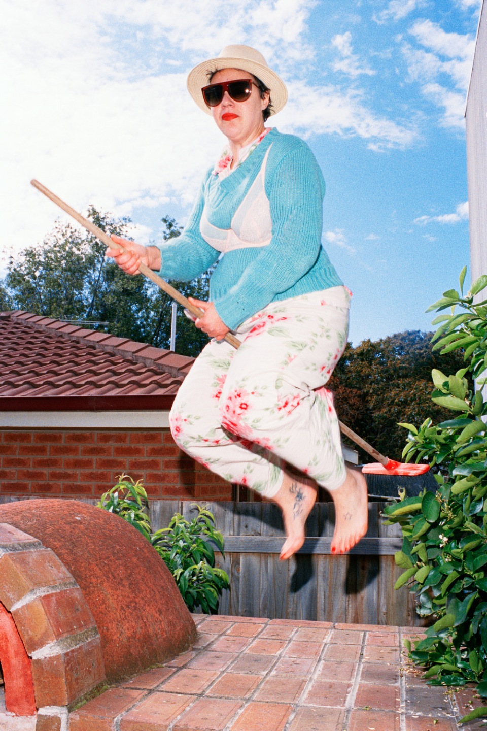 Adult female with sunglasses and sun hat jumping and holding a broomstick outdoors in front of plants and red brick oven.