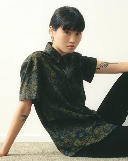 Young Asian woman in patterned shirt and black pants poses on carpet against white wall, showcasing a modern yet casual fashion style.