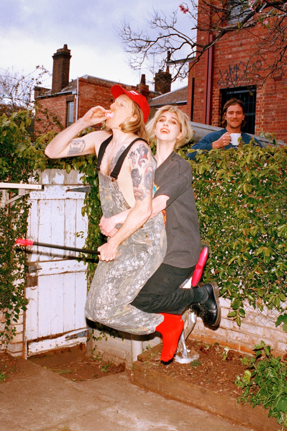 Tattooed adult male on a homemade swing, embraced by an adult female in a backyard setting, with another man in the background.