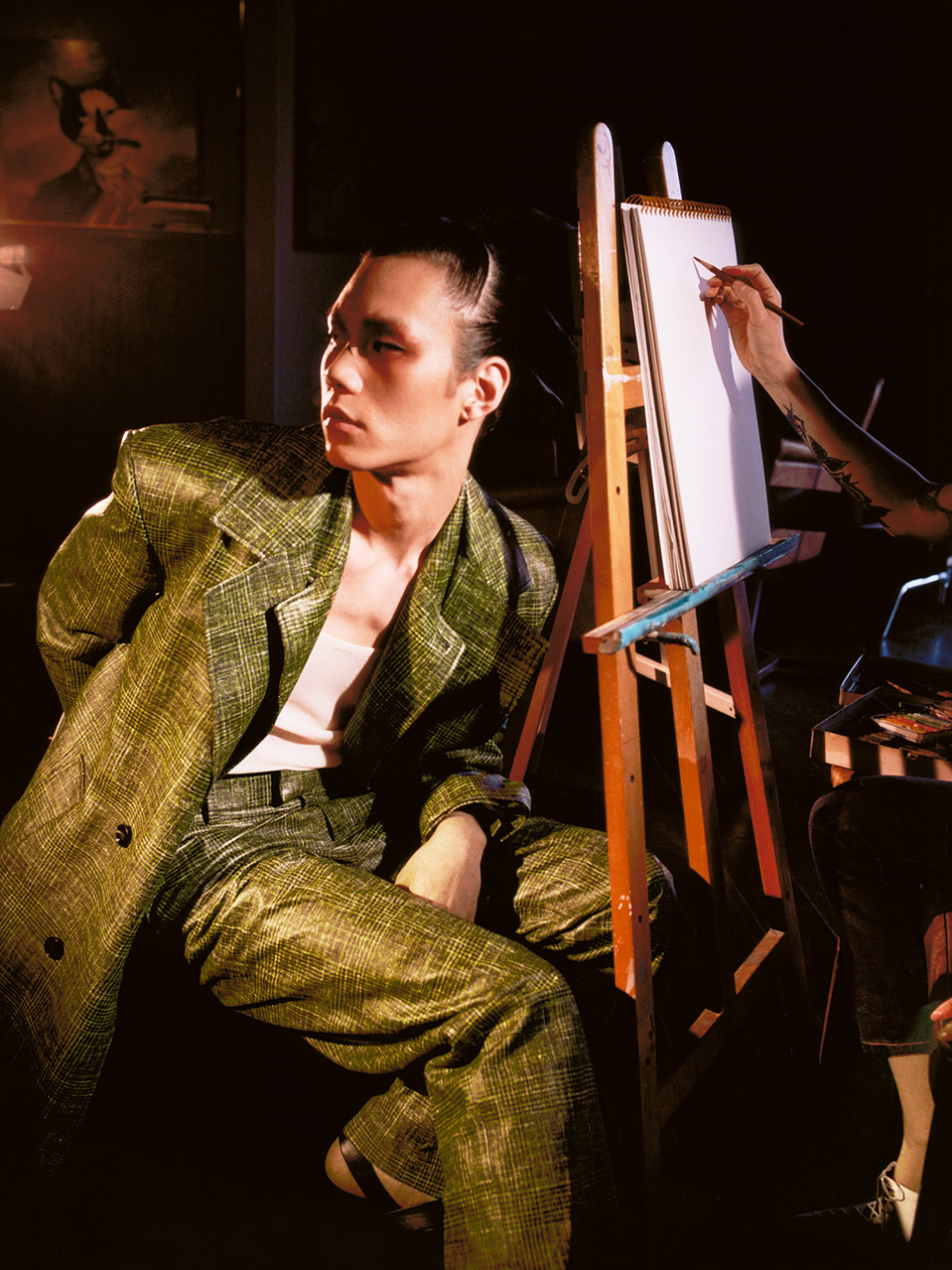 An adult male in a shiny green suit sits next to an artist's easel with a partially visible artist in a dimly lit indoor setting.