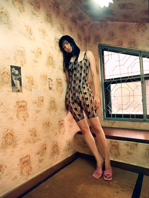 Young woman leans against patterned wall in bodysuit and pink heels, window to the side, creating a vintage-styled fashion shot.