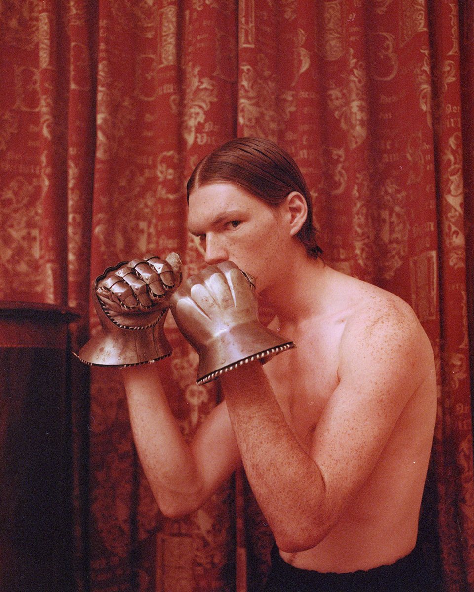An adult man with long hair in boxing pose wearing metal gauntlets in front of a red pattern curtain, medium shot, eye level angle.