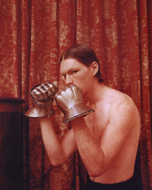 An adult man with long hair in boxing pose wearing metal gauntlets in front of a red pattern curtain, medium shot, eye level angle.