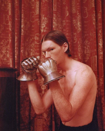 An adult man with long hair in boxing pose wearing metal gauntlets in front of a red pattern curtain, medium shot, eye level angle.