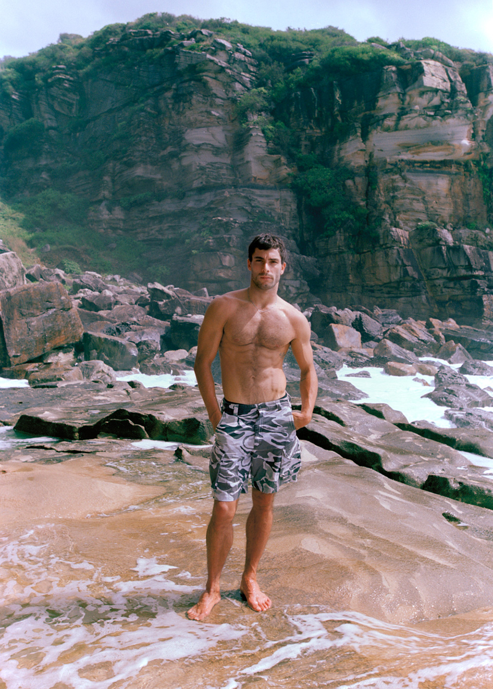 A man with a muscular build poses barefoot on a rocky beach wearing camo shorts with a mountain covered in vegetation in the background.