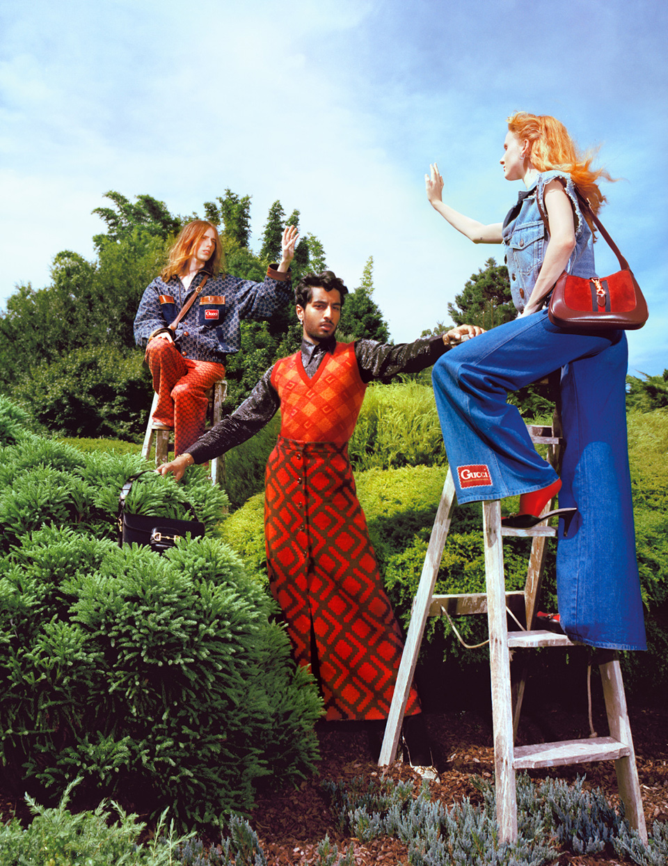 Three fashion models in vintage-inspired clothing pose on and around ladders in a lush outdoor garden setting under a bright sky.