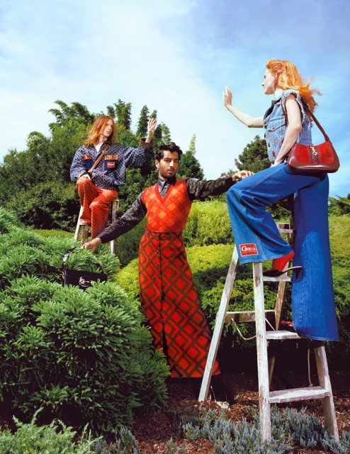 Three fashion models in vintage-inspired clothing pose on and around ladders in a lush outdoor garden setting under a bright sky.