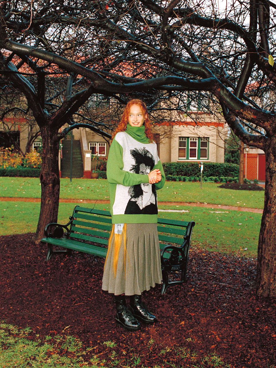 Red-haired woman in green sweater and skirt standing on mulch in a park under tree branches, with green park bench and house in background.