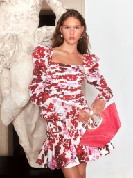 Model in a floral red and white dress standing beside a classical marble statue, holding a pink handbag.