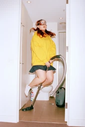 Woman wearing a yellow hoodie and face mask jumping in the air while holding a vacuum cleaner wand indoors