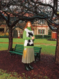 Red-haired model in a green turtleneck sweater and pleated skirt standing outdoors in a park by a green bench