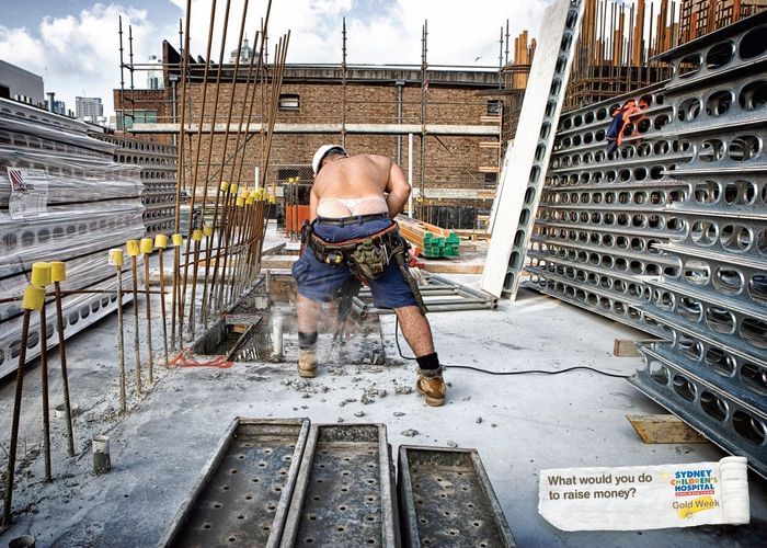 A shirtless construction worker in shorts and a hard hat is shown from behind on a building site operating a concrete saw tool.