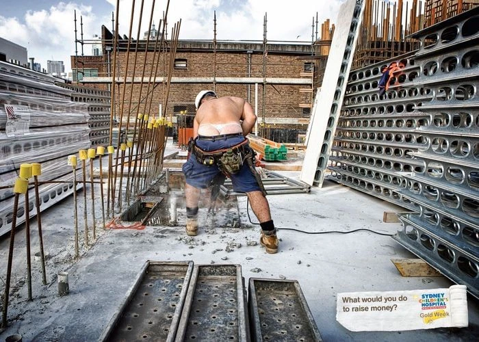 A shirtless construction worker in shorts and a hard hat is shown from behind on a building site operating a concrete saw tool.