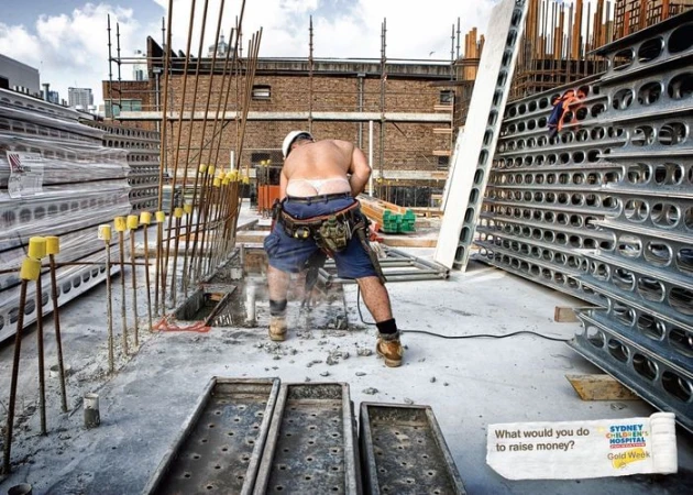 A shirtless construction worker in shorts and a hard hat is shown from behind on a building site operating a concrete saw tool.