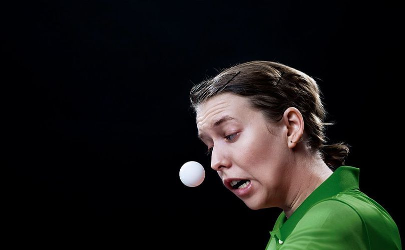 A woman in a green shirt plays table tennis, intently watching the ping pong ball against a stark black background in a close-up shot.