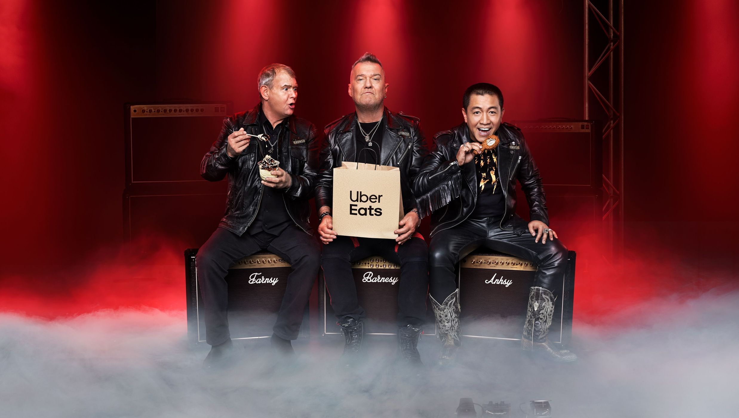 Three men in leather jackets sit on Marshall amps on stage with Uber Eats bag, dessert, and donut against a red background.