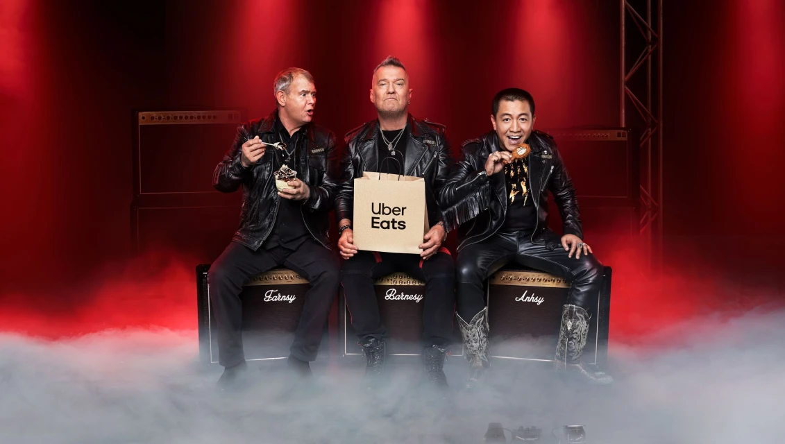 Three men in leather jackets sit on Marshall amps on stage with Uber Eats bag, dessert, and donut against a red background.