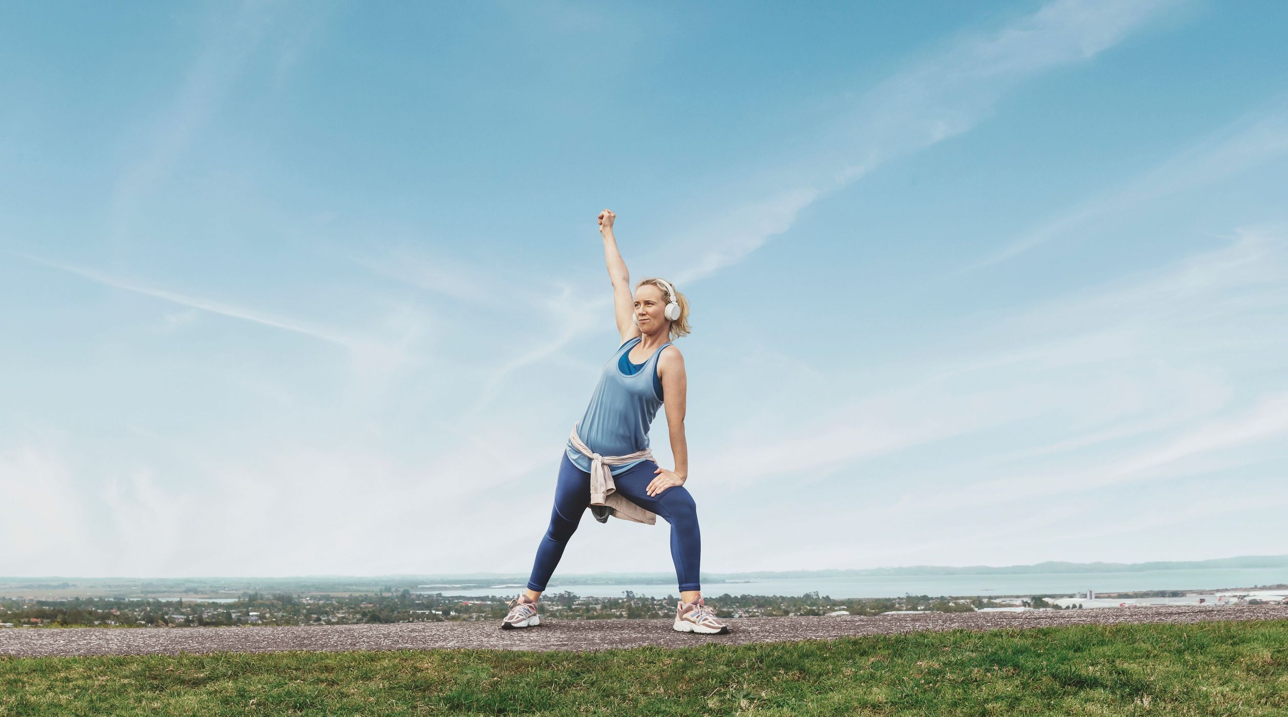 Adult woman in athletic wear stretches outdoors on a grassy hill against a blue sky with light clouds in a wide outdoor shot.