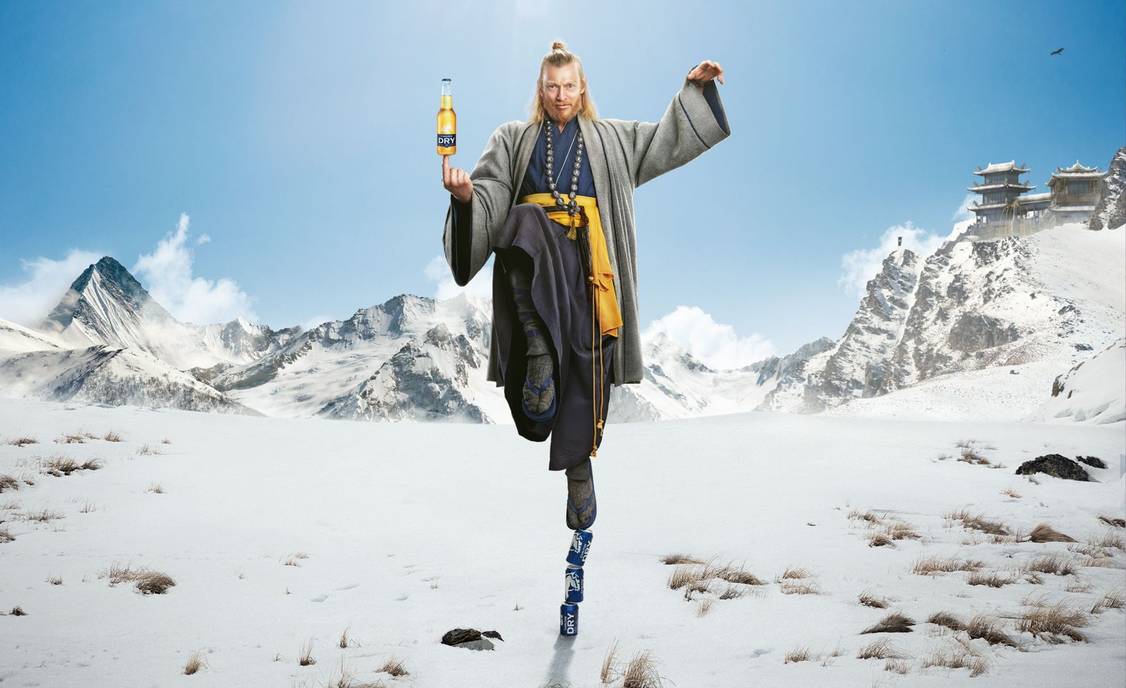 Man in monk robes balances on beer cans while holding a beer bottle in a snowy mountain landscape with temple and blue sky.