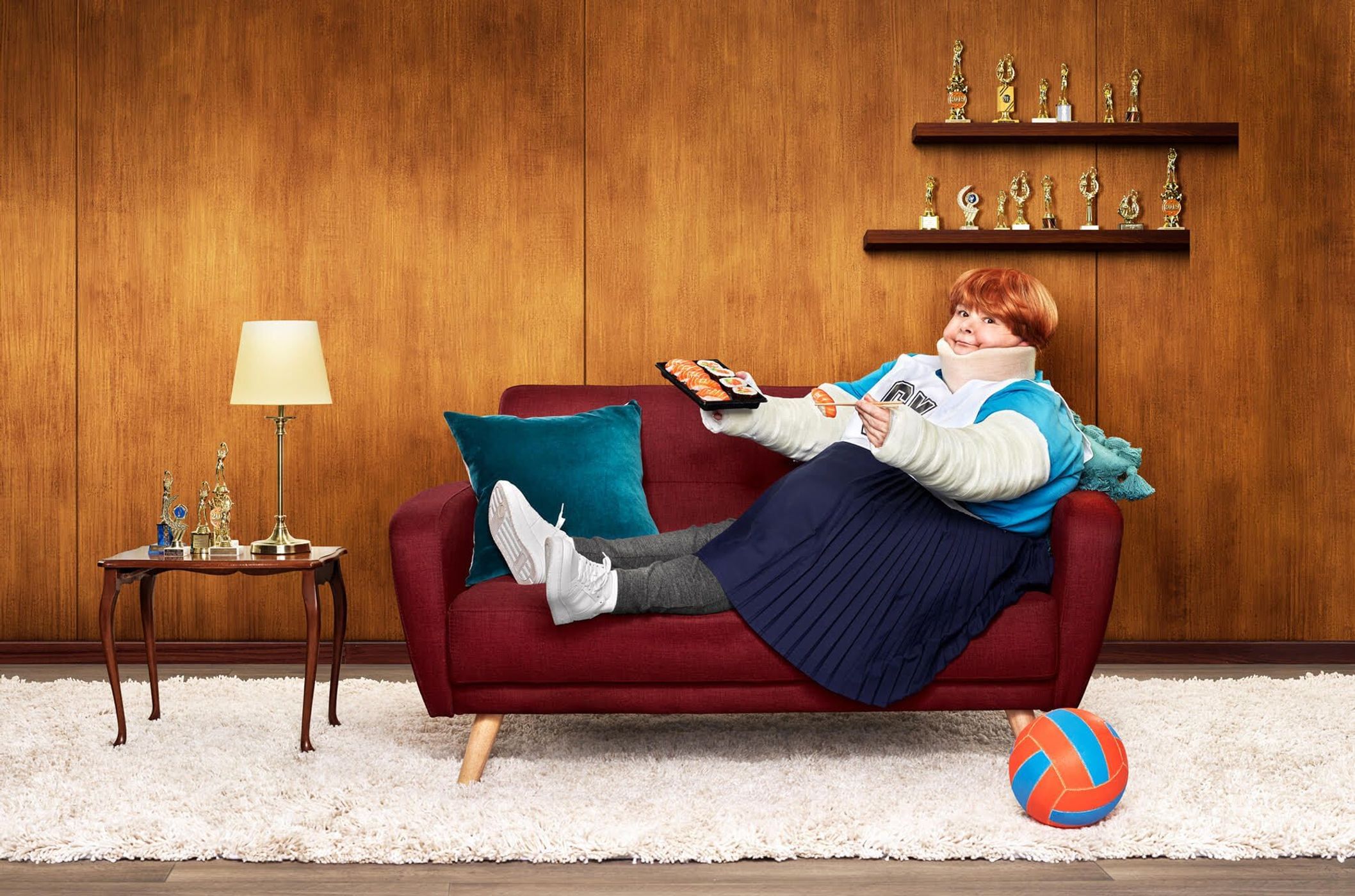 A woman with a neck brace and arm casts sits on a red couch, holding sushi, with a ball and trophies visible in an indoor setting.