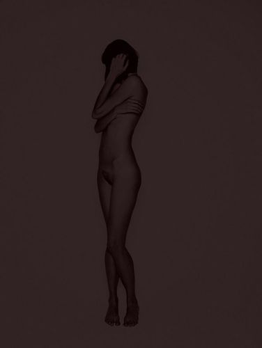 Side view of nude adult female figure with short hair in a neutral studio setting, arms crossed, standing in a dimly lit environment.