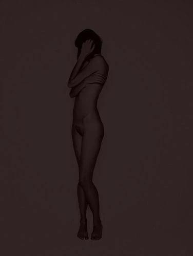 Side view of nude adult female figure with short hair in a neutral studio setting, arms crossed, standing in a dimly lit environment.