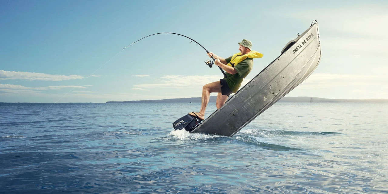 Man fishing from an angled aluminum boat in the ocean on a sunny day, wearing a life vest and hat, with blue sky and water.
