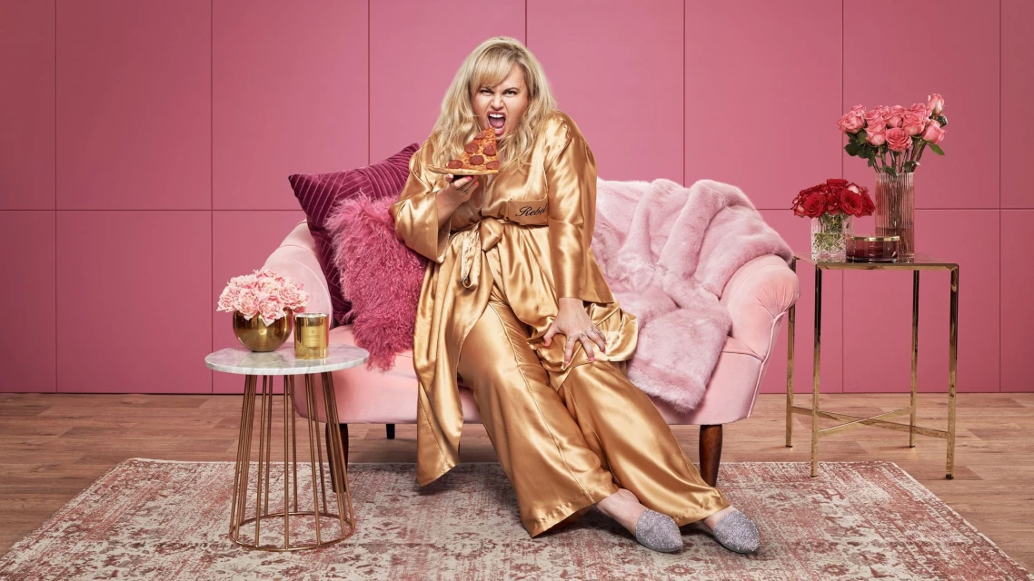 Rebel Wilson in gold pajamas eats pizza on a pink velvet couch in a photo studio setting with floral arrangements and vintage rug.