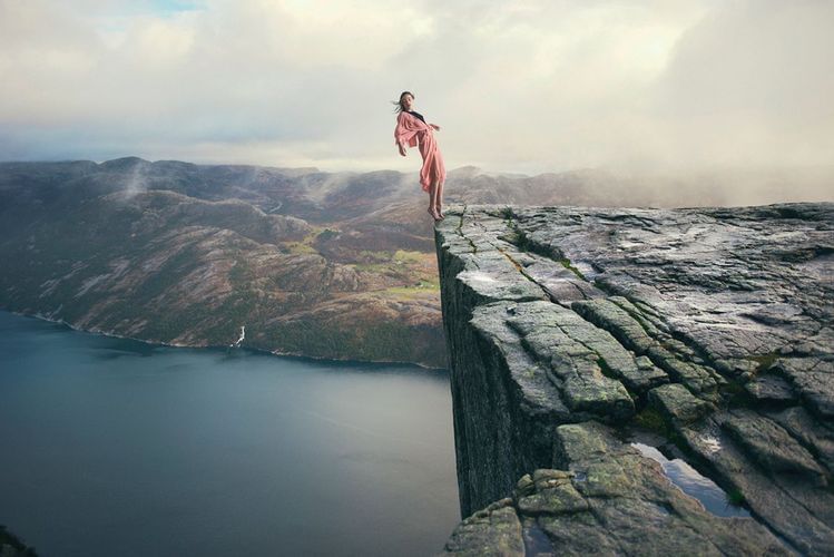 A woman in a pink robe stands on the edge of a cliff overlooking a fjord, with mountains and a cloudy sky in the background.