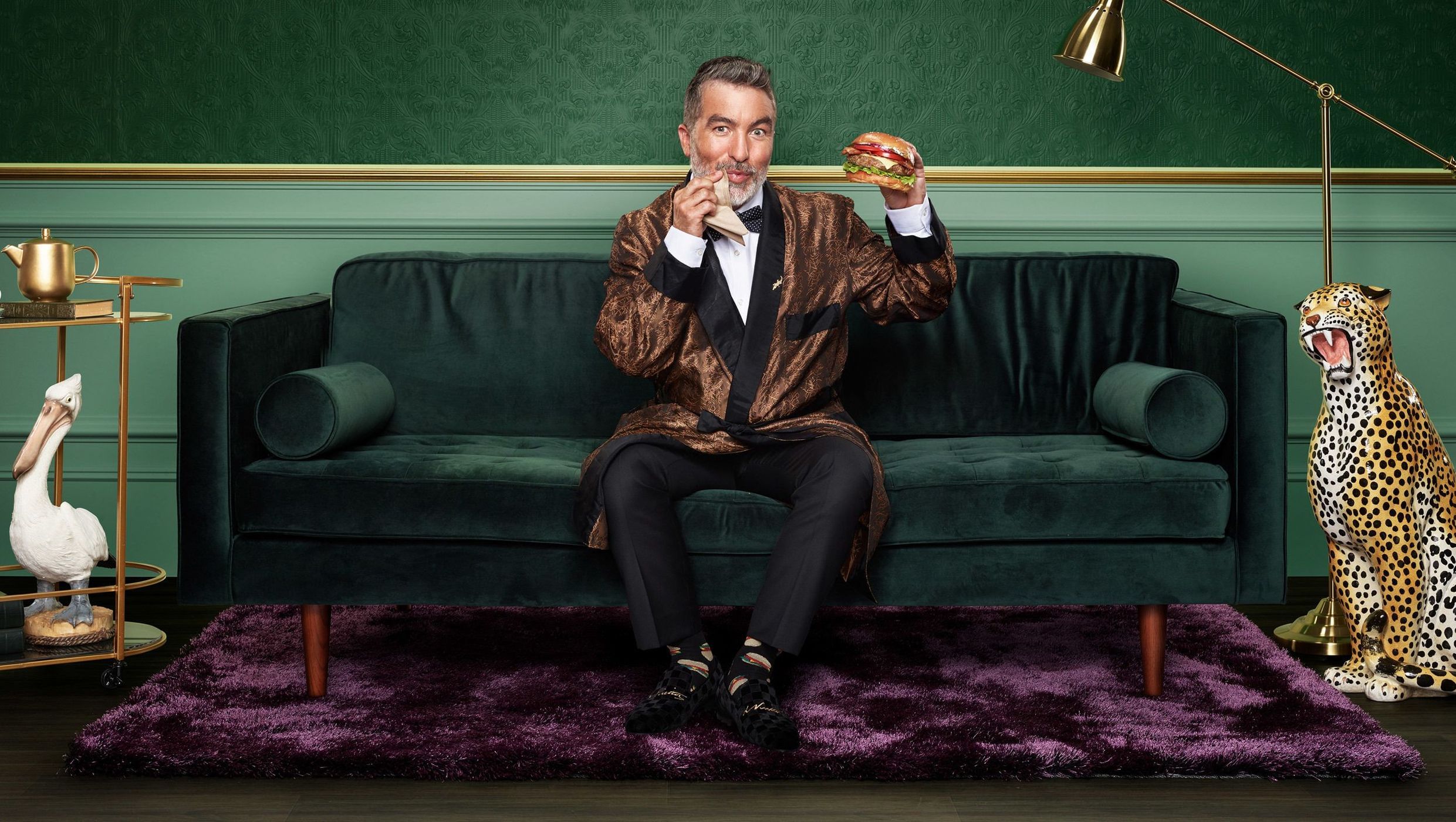 A person in a robe sits on a green velvet couch, eating a burger in an indoor setting with a faux leopard statue.